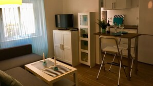 A compact living space with a sofa, a small table with candles, a television on a cabinet, a kitchen area with a table and chairs, and a dining table with wine glasses.