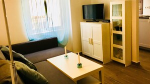A compact living space with a sofa, a television on a cabinet, a small table with a candle, and a kitchen area with cabinets and a countertop.