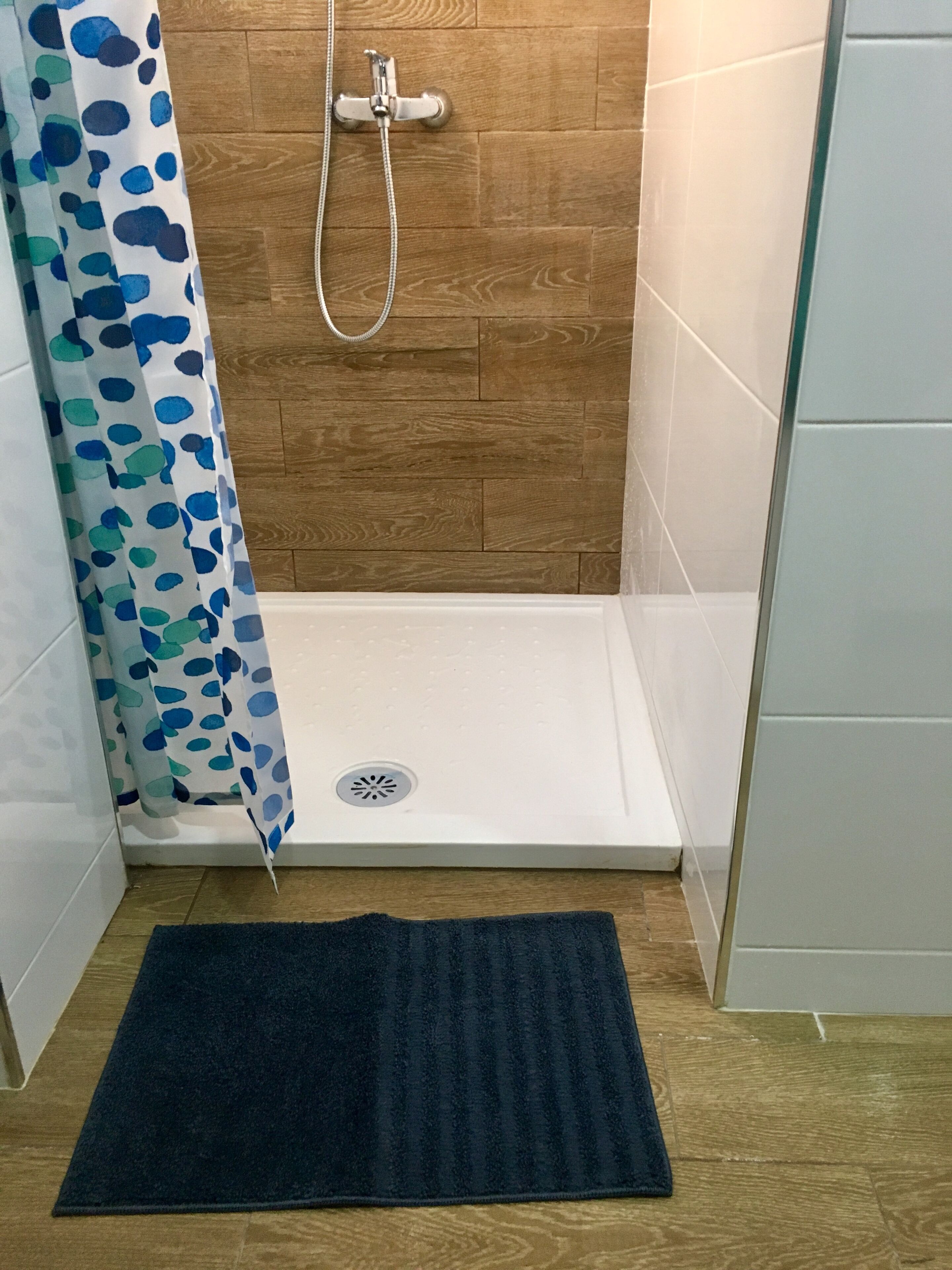 A shower area with a wooden wall, a white shower base, and a blue shower curtain with a leaf pattern.