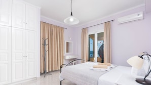 Quarto com cama, escrivaninha com abajur, guarda-roupa e banheiro visível por uma porta de vidro.