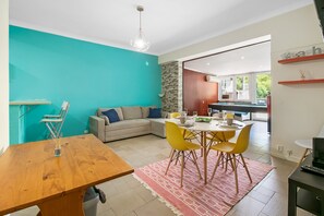 A modern living space with a teal accent wall, a wooden dining table, yellow chairs, a sofa with blue cushions, and a kitchen area visible through an open door. - CAPARICA BEACH CHARMING (Costa da Caparica)