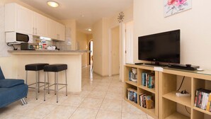 A compact living space with a kitchenette, a TV, a bookshelf, and a blue chair. - Santa Maria 88 - Two Bed (Costa Adeje)
