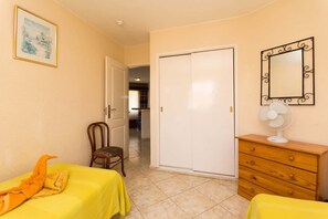 A room with two beds, a wooden dresser, a white fan, a door, and a framed picture on the wall.
