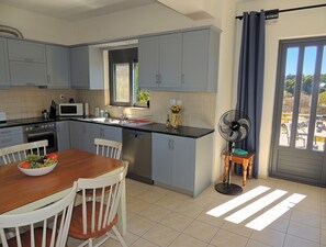 A kitchen with light blue cabinets, a wooden dining table with chairs, a microwave, and a fan near the window.