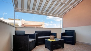 Un espace extérieur aménagé avec une zone salon couverte, doté de meubles en osier et d’une petite table, offrant une vue dégagée sur le ciel et les bâtiments en arrière-plan.