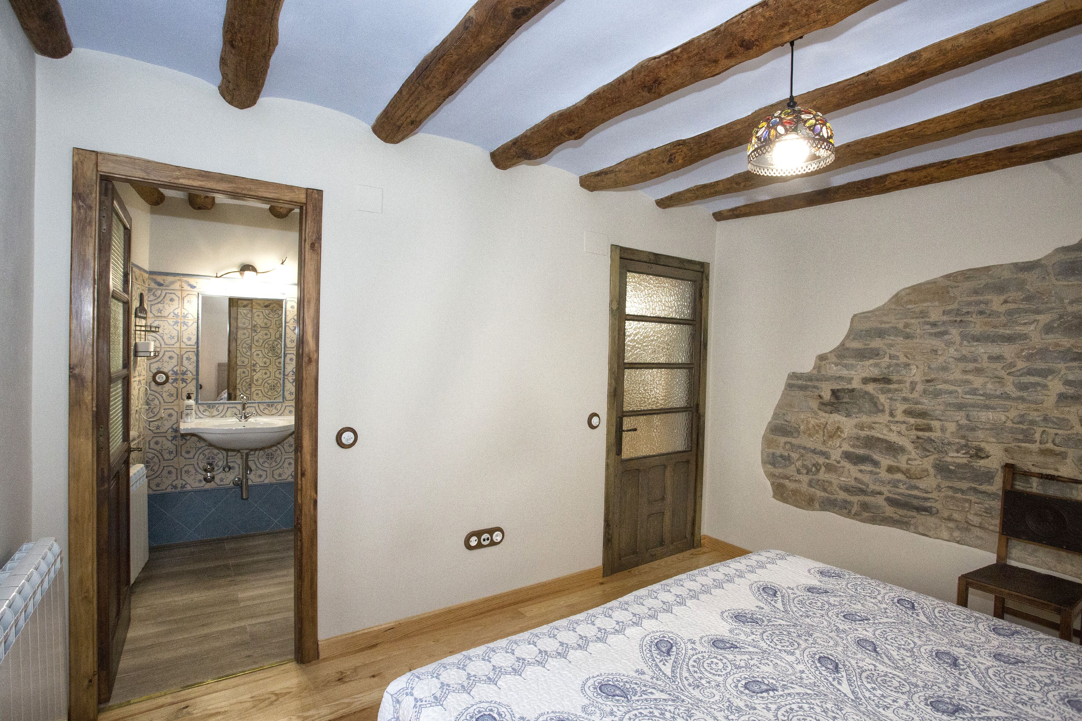 Une chambre avec un plafond en bois, un mur en pierre, un lit recouvert d’une couvre-lit à motifs, une porte en bois et une salle de bain avec un lavabo et un miroir à motifs.