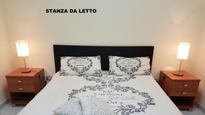 Una cama impecablemente hecha con una colcha decorativa que tiene el texto 'Eau de Cologne' y un diseño de corazón, flanqueada por dos mesitas de noche con lámparas y cajones.
