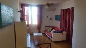 Una sala de estar con un sofá blanco, una mesita, un ventilador de techo y una ventana con cortinas.