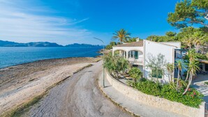Una propiedad costera con un camino pavimentado que conduce a un edificio blanco con techo de tejas, rodeado de vegetación y vistas al mar.