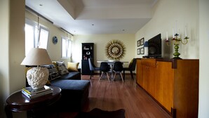 A living room with a sofa, a coffee table, a dining table with chairs, a TV, a wooden cabinet, and a decorative mirror on the wall.