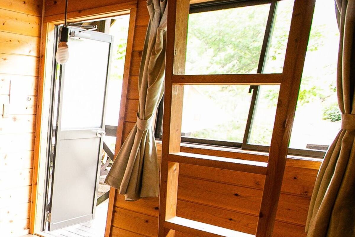 A wooden cabin interior with a sliding door leading to an outdoor area, a window with curtains, and a wooden bench.