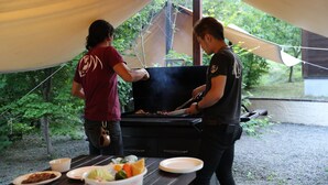Dos personas asando al aire libre bajo una carpa, con una mesa preparada con comida y platos.