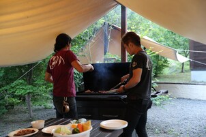 Dos personas asando al aire libre bajo una carpa, con una mesa preparada con comida y platos.