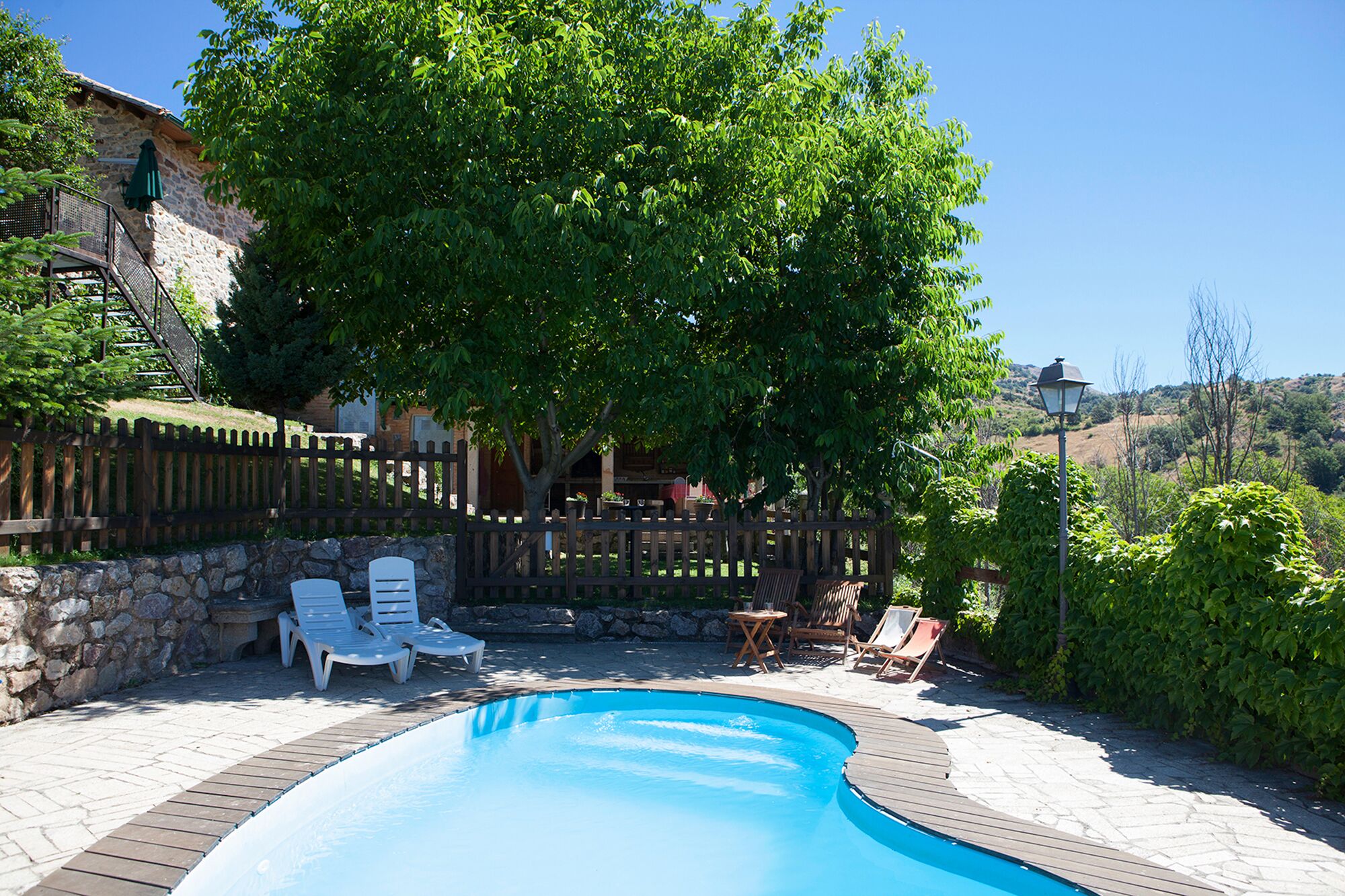 Piscina con muelle de madera y sillones de descanso, rodeada de muros de piedra y una valla de madera.