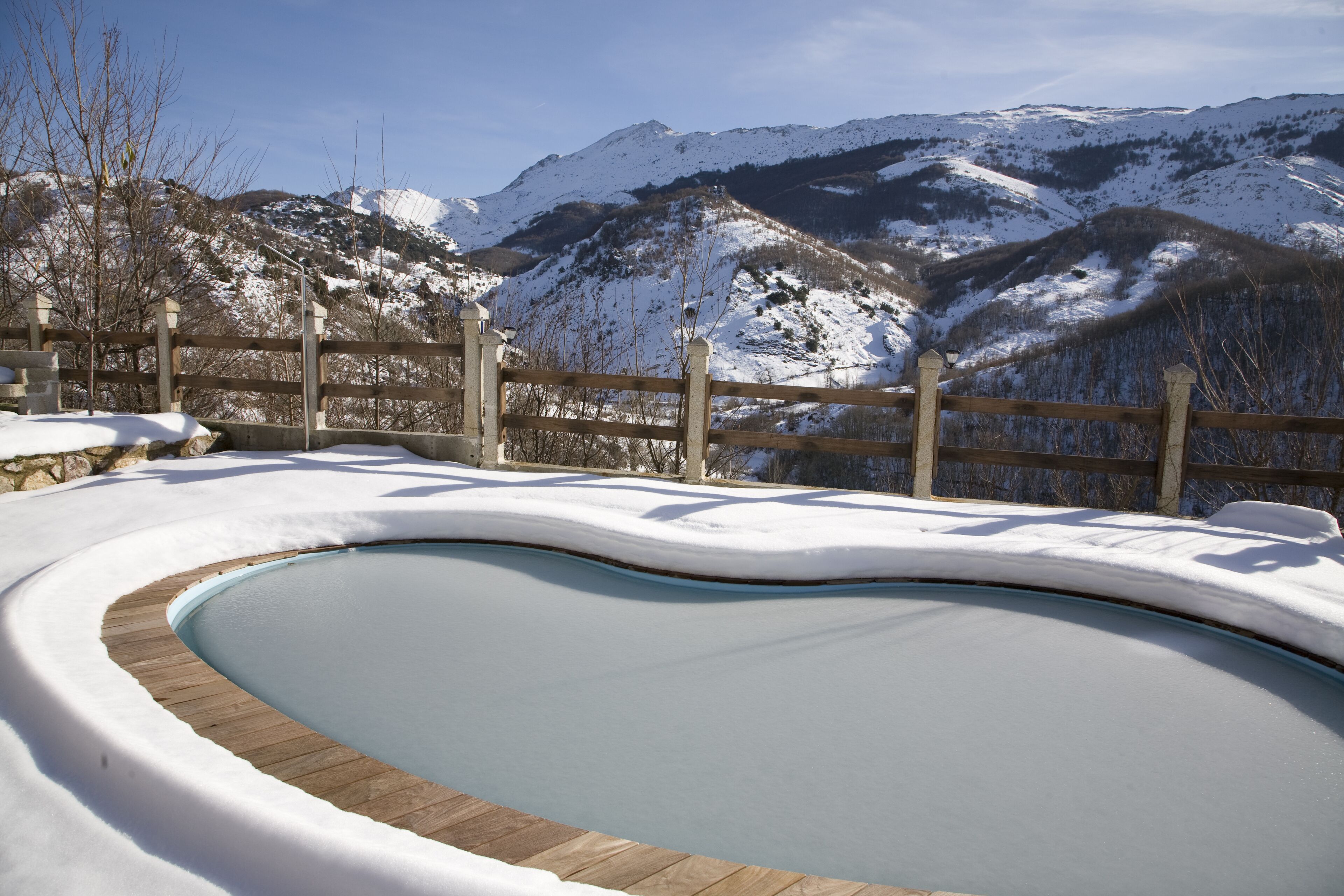Piscina exterior cubierta de nieve con un muelle de madera, rodeada por una valla de madera y montañas al fondo.