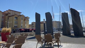 Outdoor seating with wicker chairs and tables, a clear sky, and buildings with balconies in the background. - Studio Gruissan plage wifi piscine, clim, parking, équipée 4 pers, RDC, terrasse sud (Gruissan Plage)