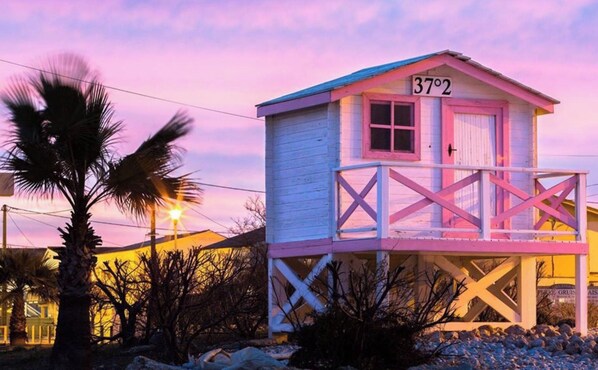 Une maison de plage avec un balcon, une pancarte indiquant « 37°2 » et un palmier au premier plan.