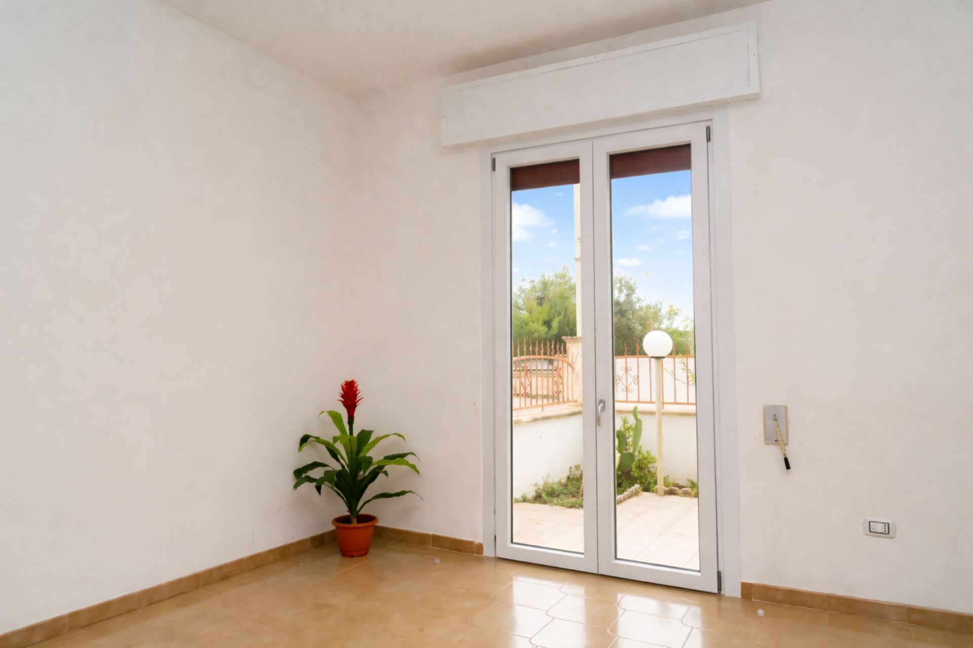 Una stanza con una porta scorrevole in vetro che dà su uno spazio esterno, una pianta in vaso e una presa elettrica sulla parete.