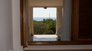 A room with a window overlooking a balcony with a view of greenery and a distant structure. - Veranda Piau  (Tertenia)