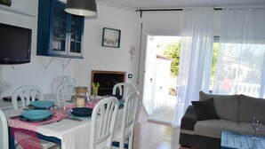 Comedor con una mesa preparada para cuatro personas, sillas blancas, un televisor montado en la pared y vistas al exterior a través de una puerta.