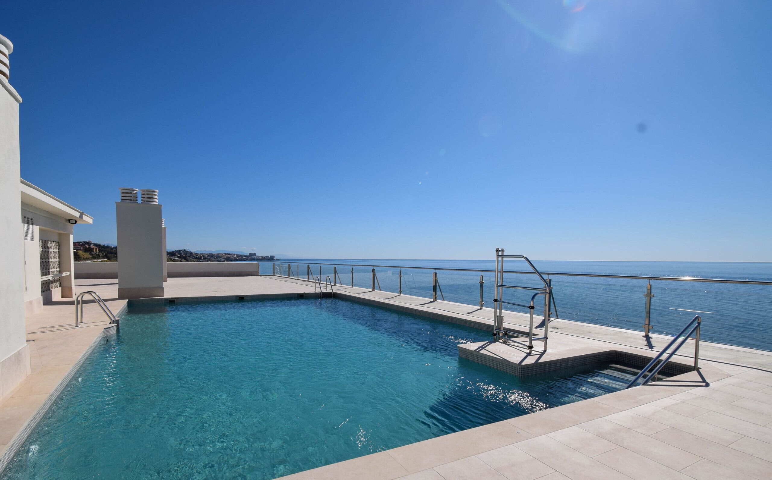 En rektangulär swimmingpool med en metallstege och räcken, belägen intill en byggnad med en vit fasad och fri utsikt över havet.