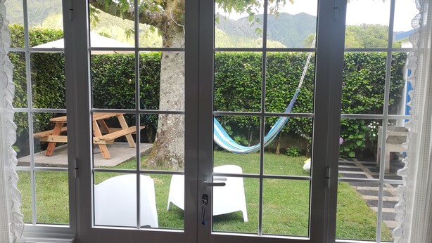 Vista a través de una ventana con una mesa y sillas de madera en el exterior, una hamaca y un área con césped y un árbol.
