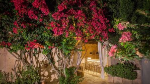 Een buitenterras met een stenen vloer, een houten pergola bedekt met roze bougainvillea, en een bar met krukken en een aanrecht.