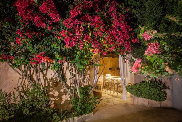 An outdoor patio area with a stone floor, a wooden pergola covered in pink bougainvillea, and a bar area with stools and a counter.