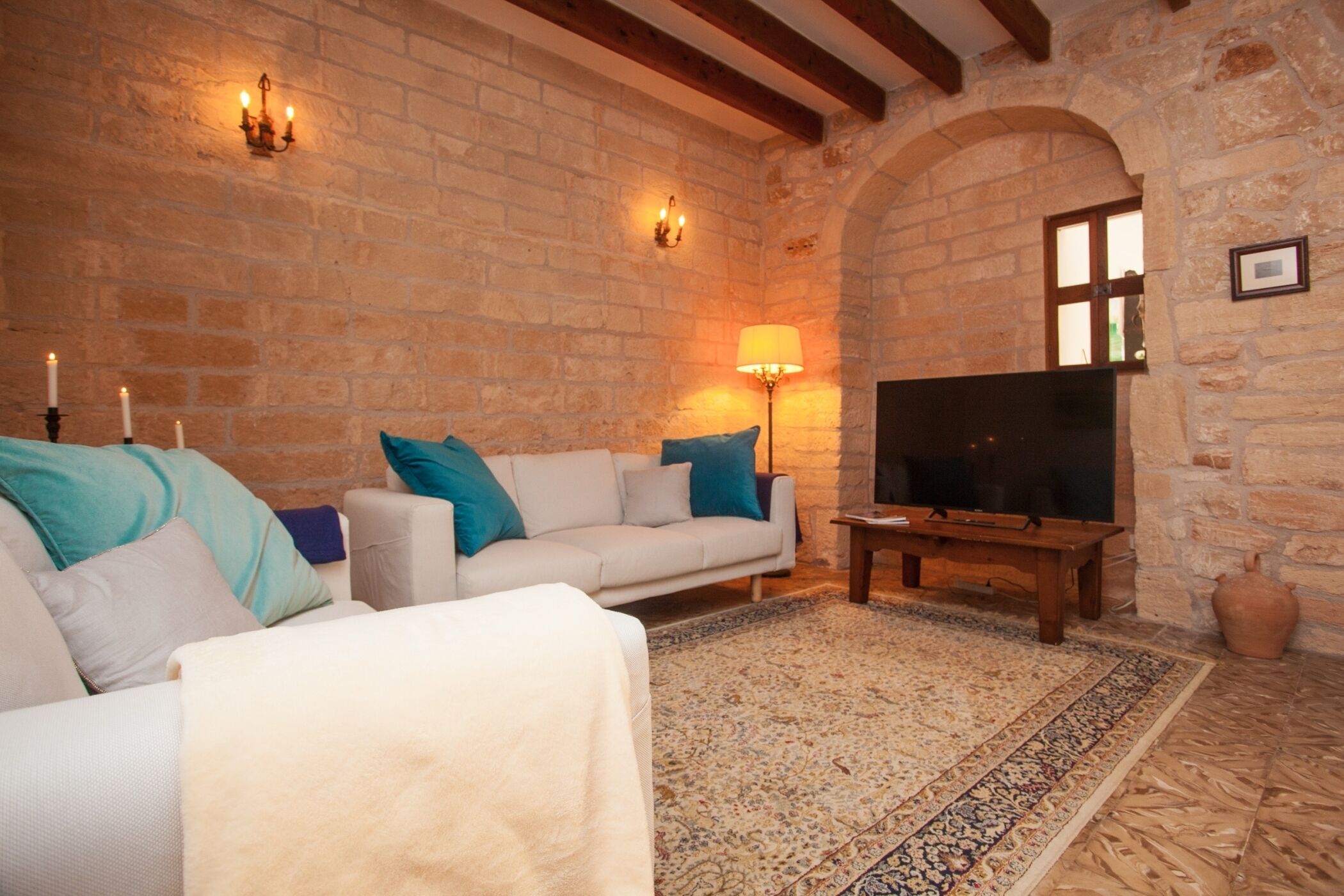 A cozy living room with a stone wall, a white sofa with blue cushions, a wooden coffee table, and a flat-screen TV.