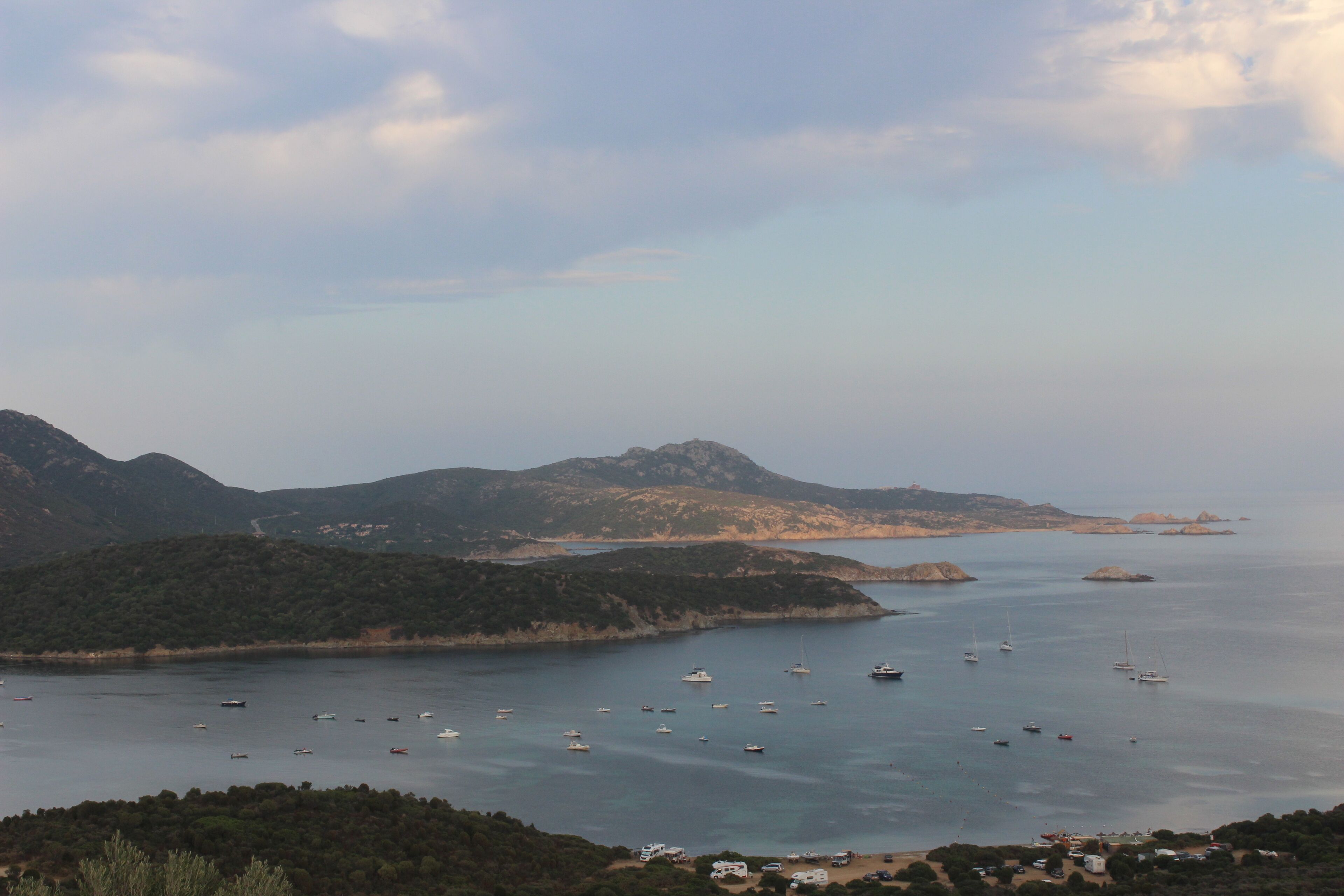 A coastal landscape with boats on the water, mountains in the background, and a small settlement with buildings and greenery in the foreground.