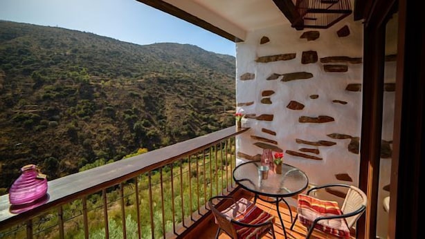 A balcony with a stone wall, a table with chairs, and a view of a hilly landscape.