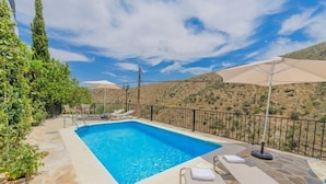 A rectangular swimming pool with clear blue water, surrounded by stone tiles and a metal fence. There are lounge chairs and umbrellas along the poolside.