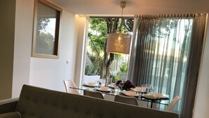 A dining area with a glass table set for four, a sofa in the foreground, and a view of greenery outside.