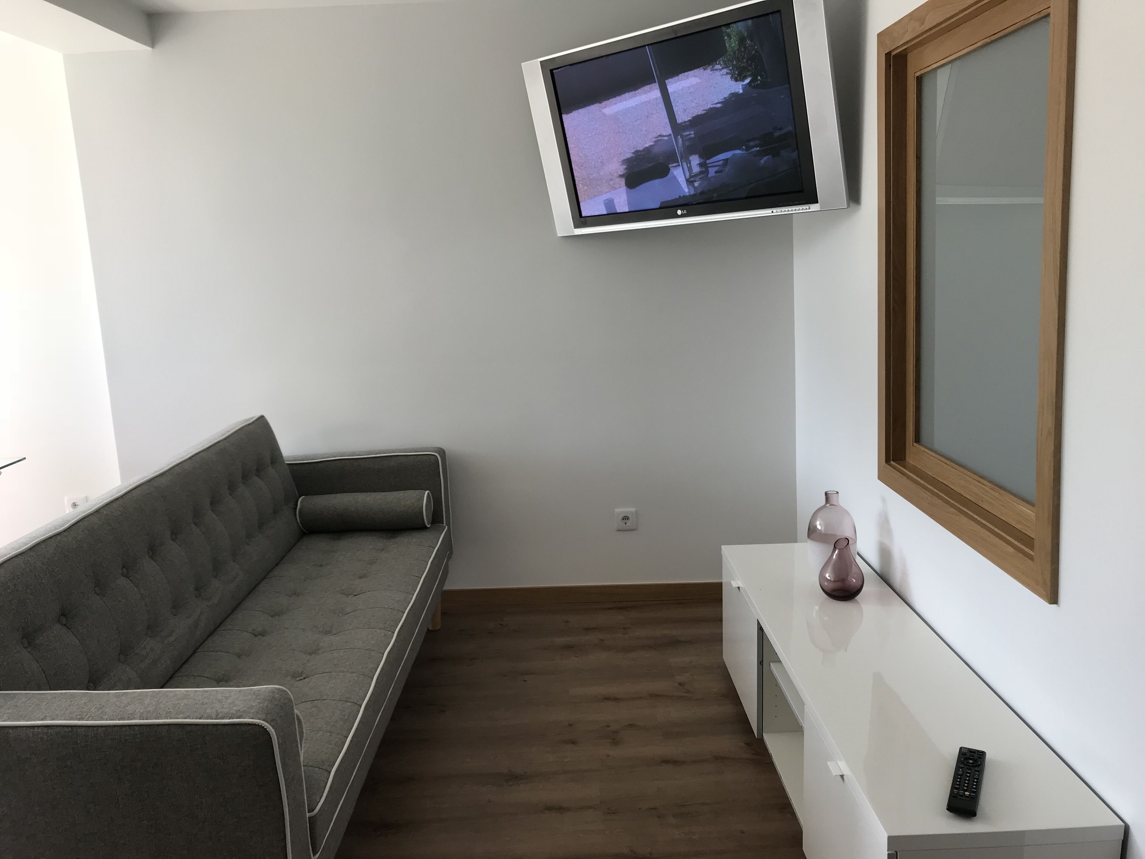 A modern living room with a grey sofa, a mounted TV, a wooden-framed window, and a white console table with a vase and remote control.