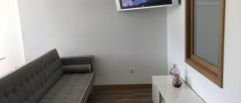 A modern living room with a grey sofa, a mounted TV, a wooden-framed window, and a white console table with a vase and remote control.