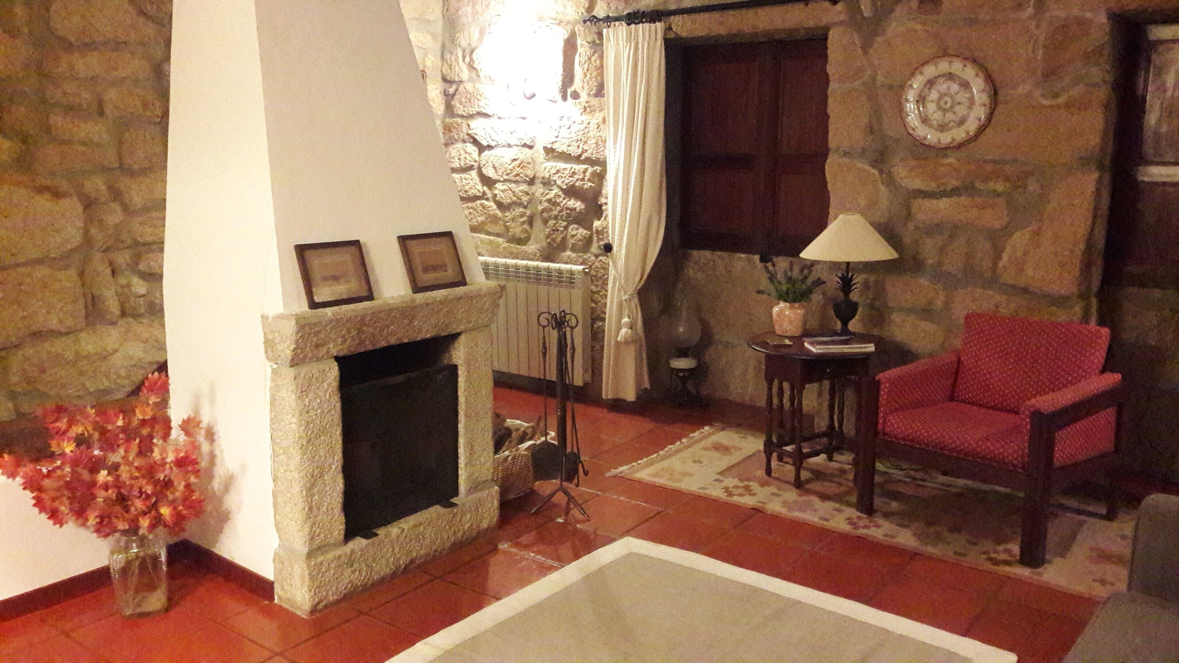 Ein gemütliches Wohnzimmer mit Kamin aus Stein, einem roten Sessel und einem kleinen Tisch mit Lampe.
