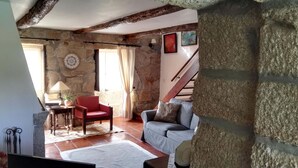 Ein rustikaler Raum mit freiliegenden Steinwänden, einer Holzbalkendecke, einem roten Sessel, einem grauen Sofa und einer Treppe, die in einen oberen Bereich führt.