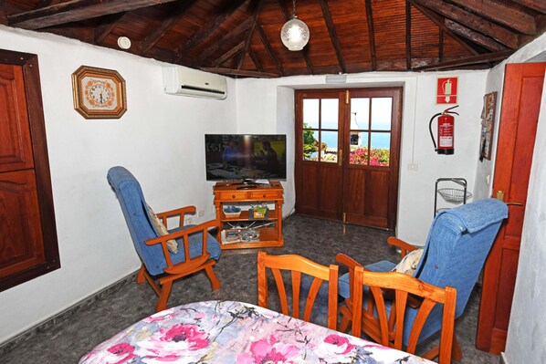 A living room with a television, wooden chairs, a table with a floral tablecloth, and a view through a door.