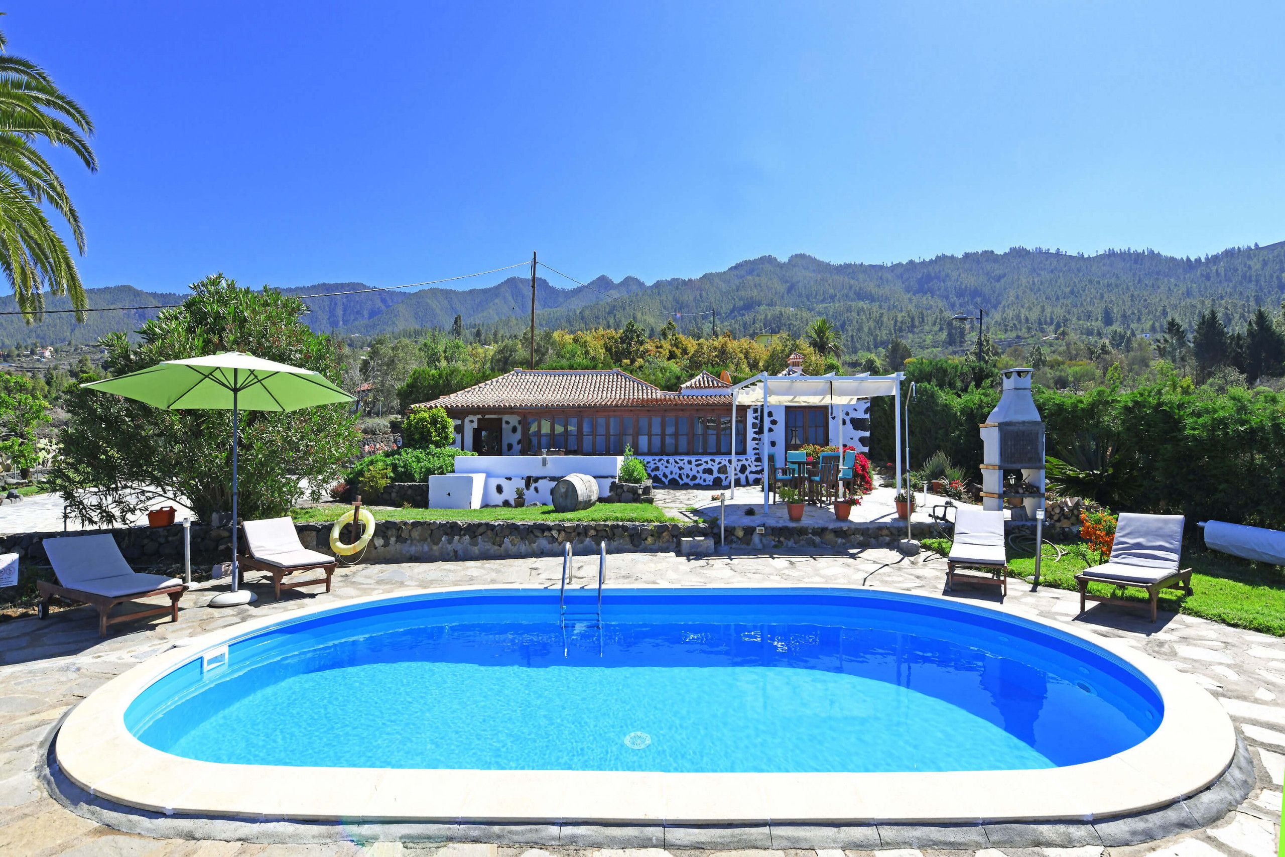 A swimming pool with lounge chairs and a green umbrella, surrounded by a stone deck and a house with a tiled roof.