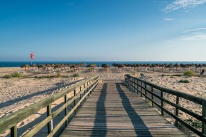 Ein Holzsteg führt zu einem Strand mit Reetsonnenstühlen und einem Rettungsschwimmerstand.