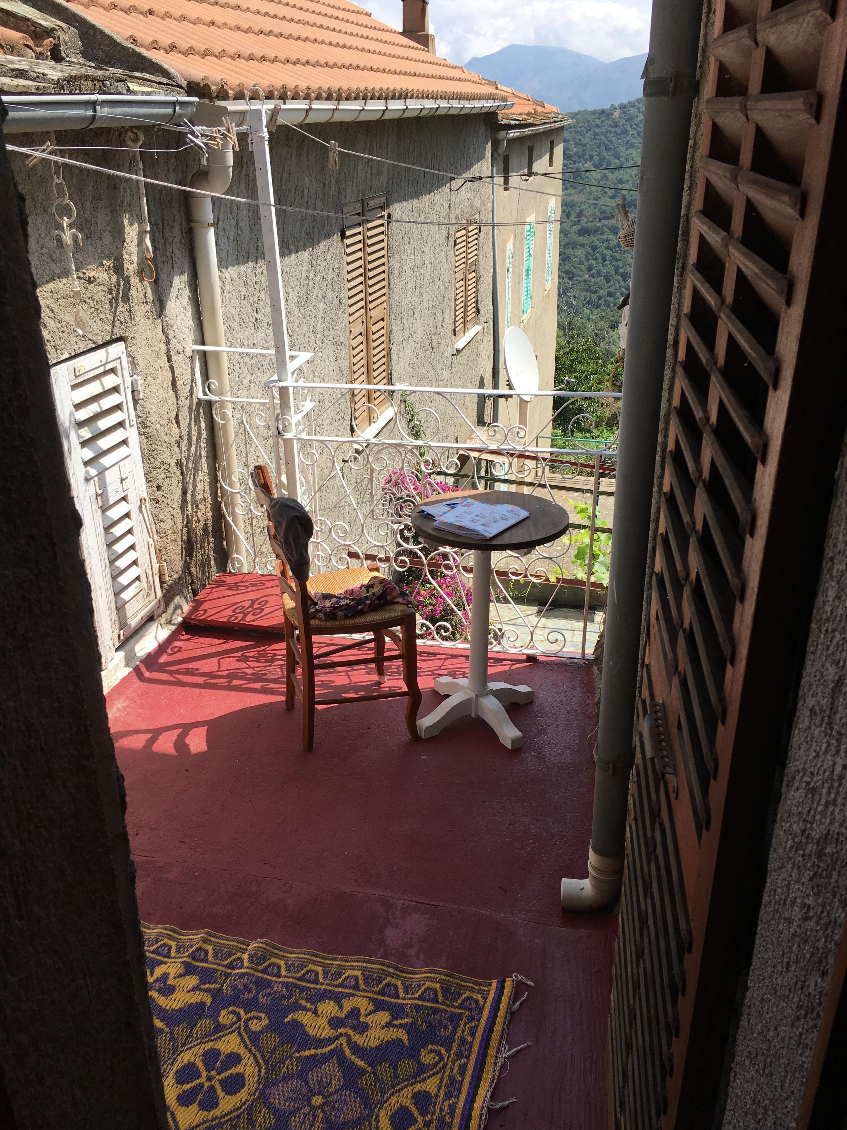 Un balcon doté d’un sol rouge, d’une chaise en bois, d’une petite table ronde sur laquelle se trouve un carnet, et qui offre une vue sur un paysage montagneux.