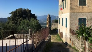 A narrow street with a stone wall on one side and a building with green shutters. A tree and mountains are visible in the background. - Village house in the heart of Sorio (Sorio)