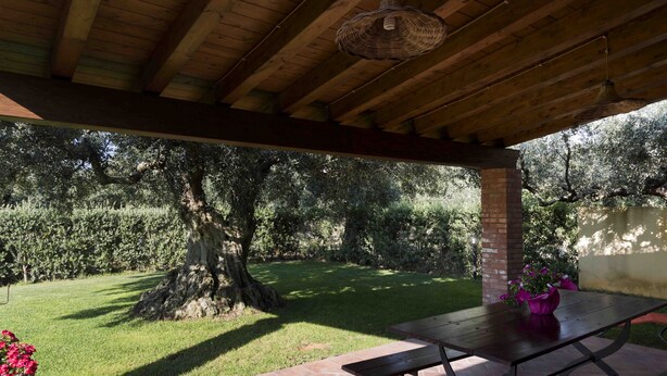 Een overdekt terras met een houten plafond, een bakstenen pilaar en een tafel met een bloevas.