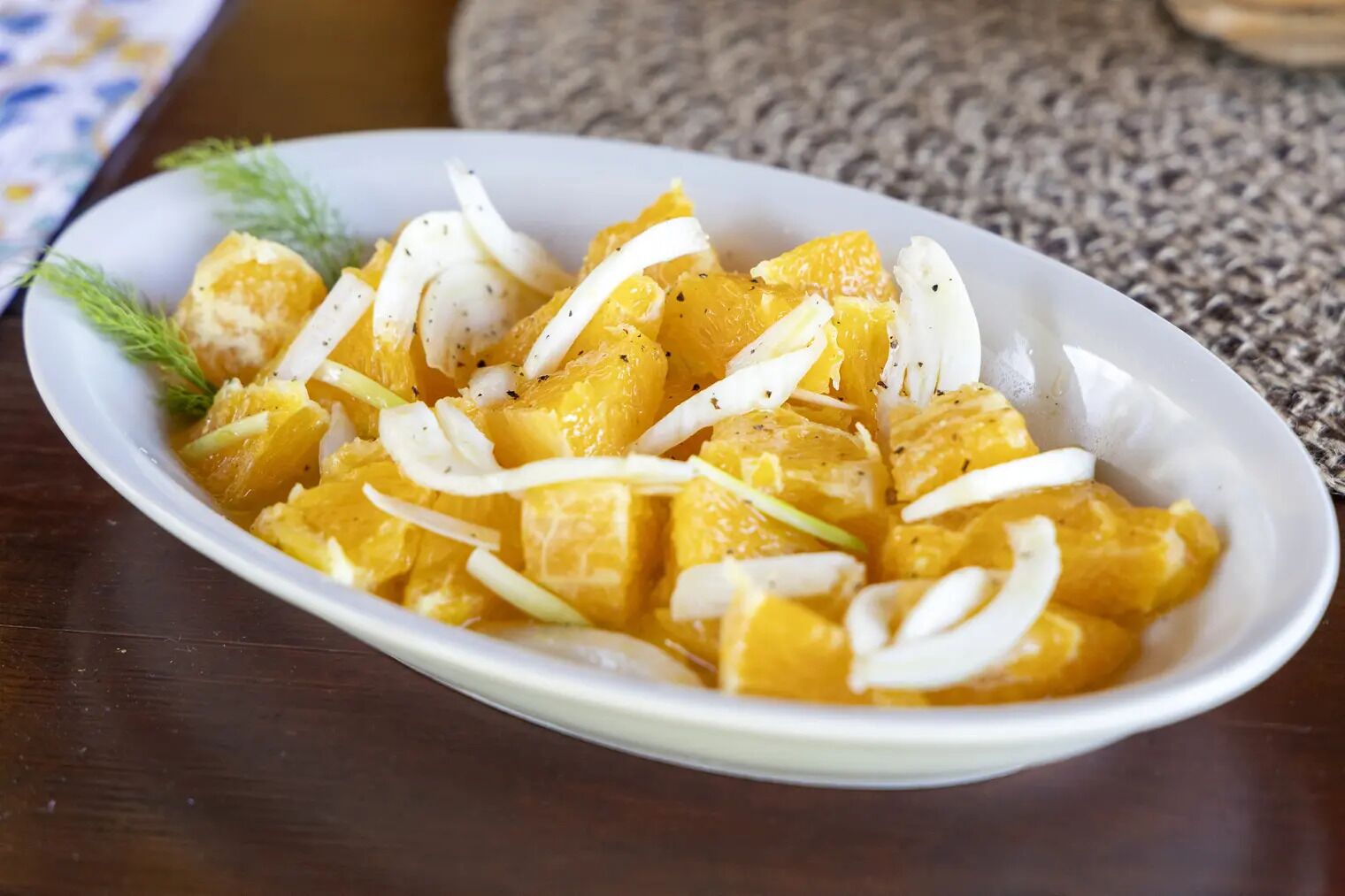 A white oval plate with cubed orange fruit, possibly mango, garnished with thin white slices and a sprig of dill.