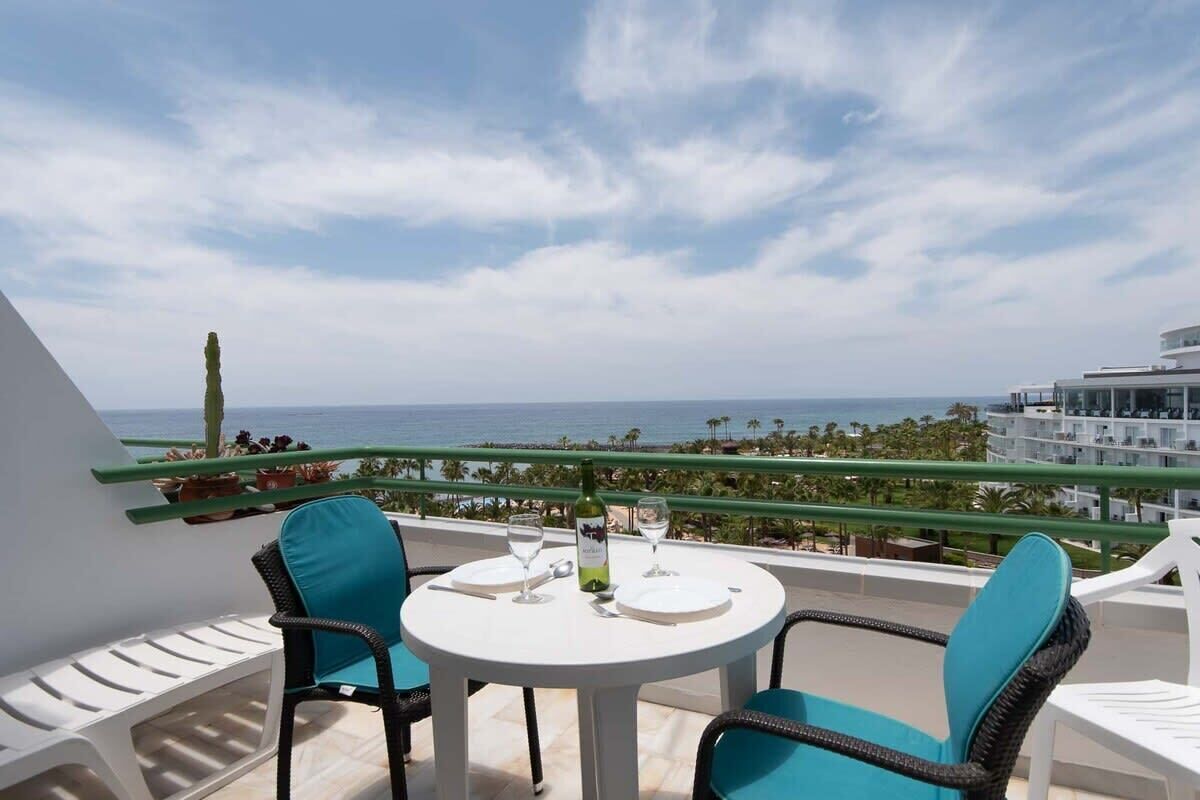 Un balcon avec une table blanche et deux chaises, d’où l’on peut admirer une vue sur la côte, avec des palmiers et un bâtiment qui se dessine à l’horizon.