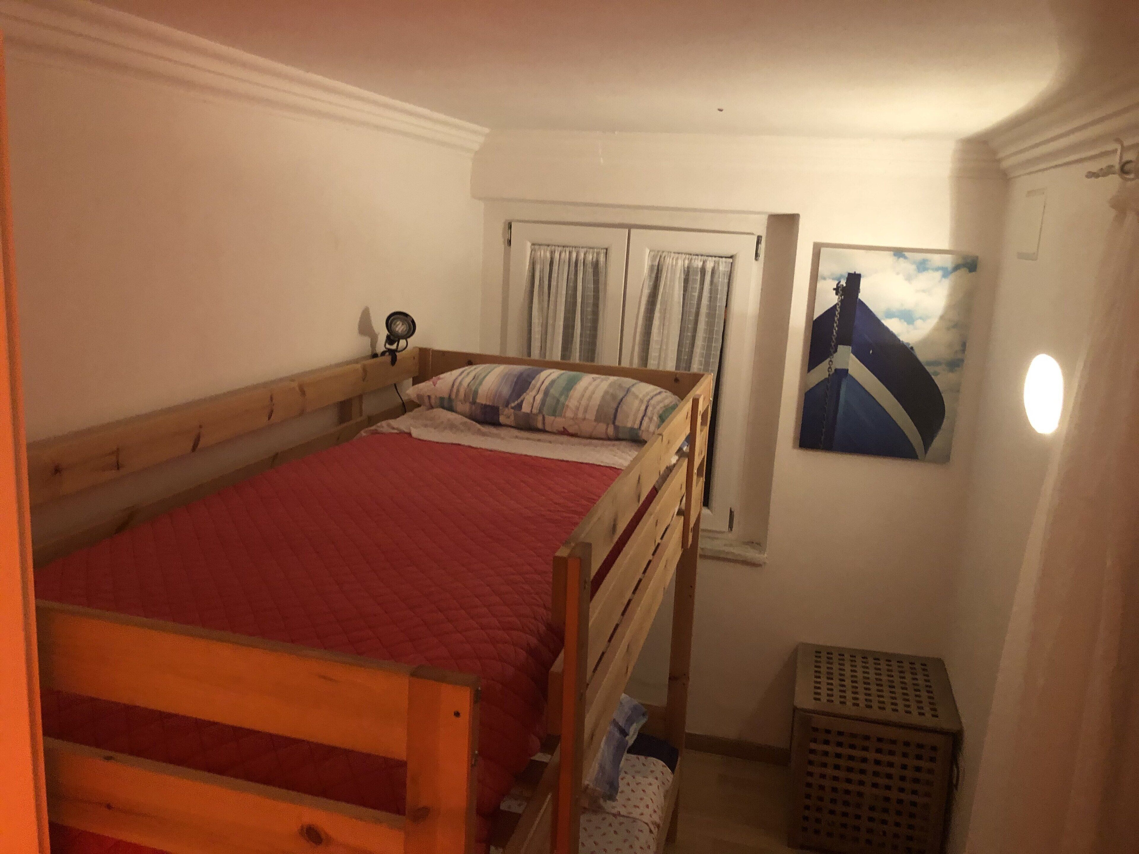 A single bed with a wooden frame and a red quilt, a small bedside table with a lamp, a window with curtains, and a wall-mounted picture.