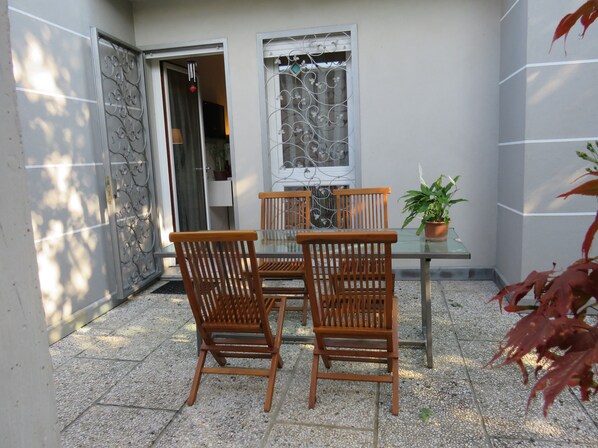 Eine Terrasse mit einem Glastisch, Holzstühlen, einem Blumentopf und einem dekorativen Fenstergitter.