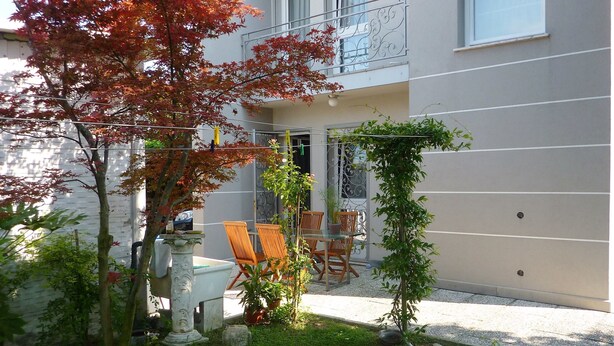 Un patio con una mesa y sillas, rodeado de plantas y un árbol pequeño con hojas rojas.