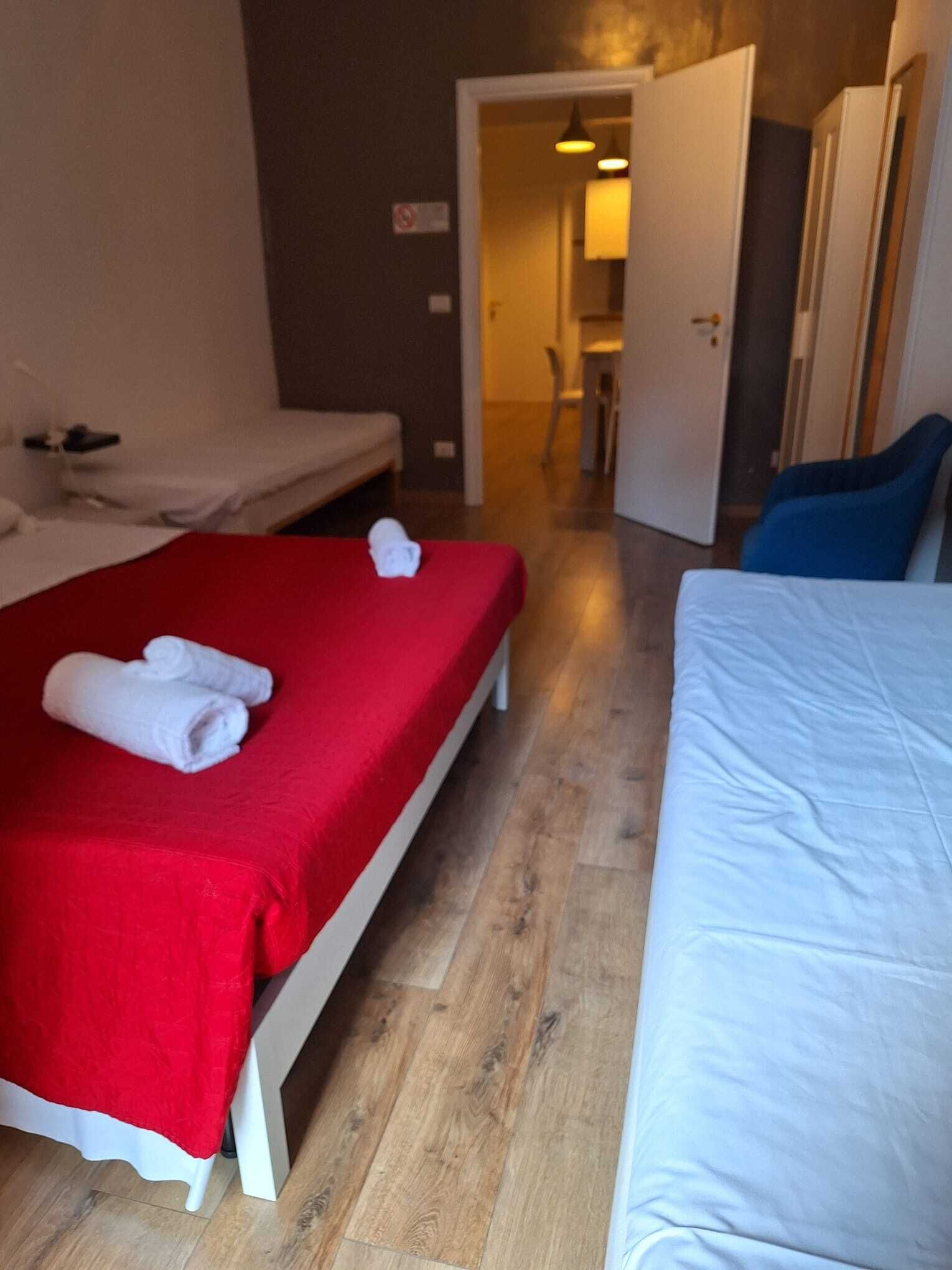 Une chambre d’hôtel avec deux lits, l’un recouvert d’une courtepointe rouge et l’autre d’une courtepointe blanche. Il y a des serviettes sur les lits, et la chambre est dotée d’un plancher en bois.