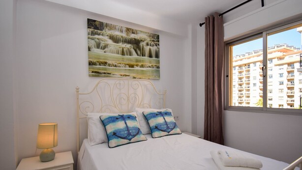 Ein Schlafzimmer mit einem Bett, Kissen, einem Nachttisch, einer Lampe, einem Fenster und einem Bild an der Wand.